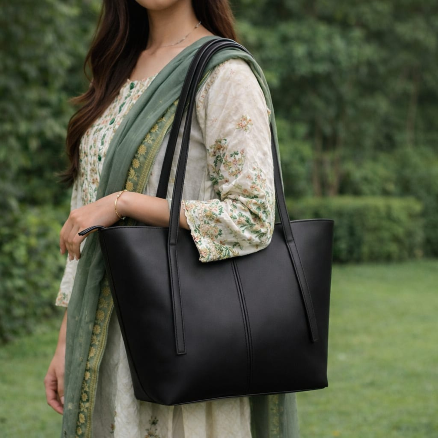 Woman holding a black leather tote bag outdoors with greenery in the background