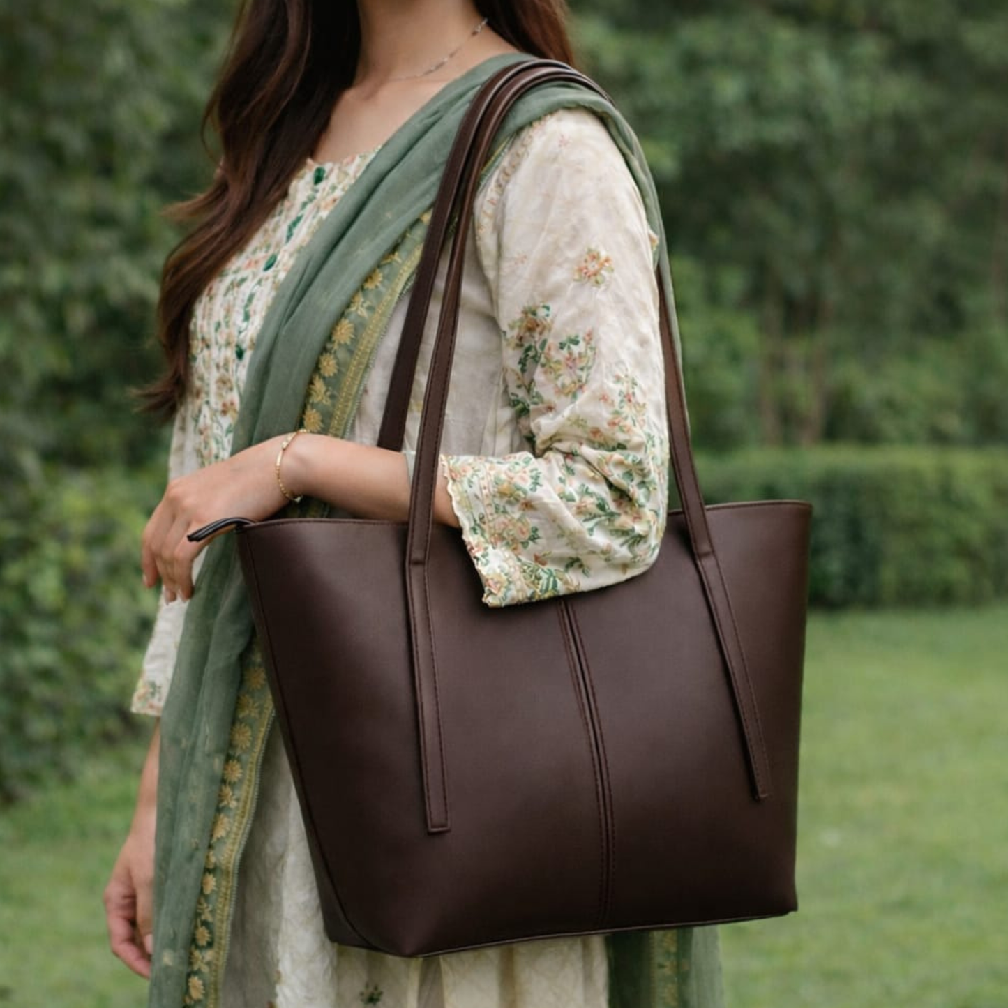 Woman holding a brown leather tote bag outdoors with greenery in the background